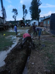 Lots going on.  Drilling, water flowing, tank stand in background being welded, trenches for piping.